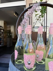 Bergischer Weinkeller Rosa etikettierte Weinflaschen stehen in einer offenen Holztür unter einer blühenden Dekoration.
