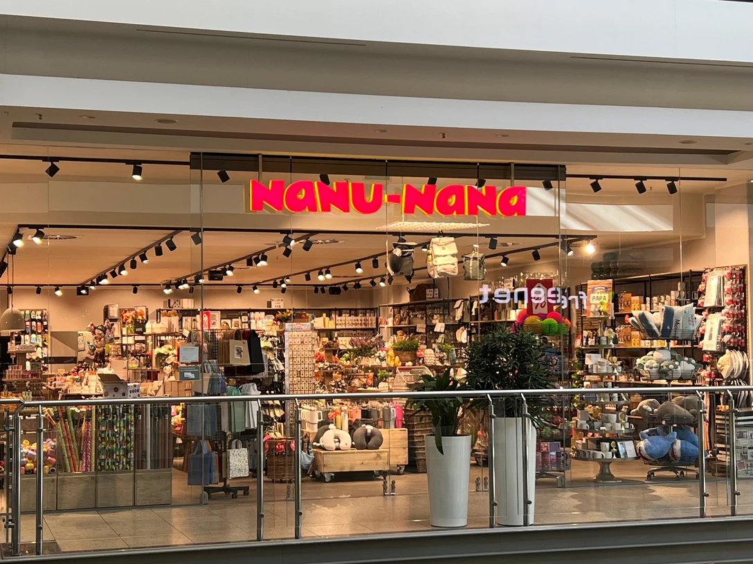 Nanu Nana Das Bild zeigt den Eingang eines Nanu-Nana-Geschäfts in einem Einkaufszentrum. Durch die gläserne Fassade sind viele bunte und dekorative Artikel zu sehen, die im Inneren des Ladens auf Regalen ausgestellt sind.