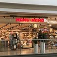 Nanu Nana Das Bild zeigt den Eingang eines Nanu-Nana-Geschäfts in einem Einkaufszentrum. Durch die gläserne Fassade sind viele bunte und dekorative Artikel zu sehen, die im Inneren des Ladens auf Regalen ausgestellt sind.