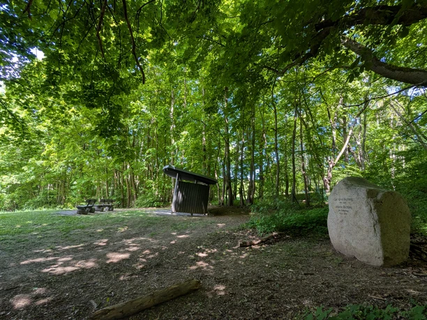 Gedenksteen met inscriptie in het bos, naast een afdak en banken onder een dicht bladerdak.