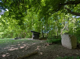 Büchting Gedenkstein_Schutzhütte_Ausblick Gedenkstein mit Inschrift im Wald, daneben Schutzhütte und Sitzbänke unter dichtem Blätterdach.