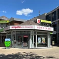 Amplifon.jpg Das Bild zeigt ein modernes Geschäft von Amplifon, spezialisiert auf Hörgeräte, mit auffälliger, roter Beschilderung und großflächigen Fenstern.