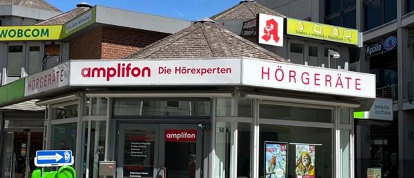 Amplifon.jpg Das Bild zeigt ein modernes Geschäft von Amplifon, spezialisiert auf Hörgeräte, mit auffälliger, roter Beschilderung und großflächigen Fenstern.