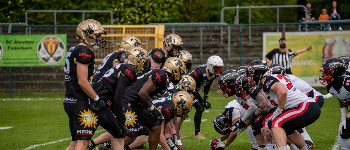 Paderborn Dolphins Football.jpg Zwei American-Football-Teams stehen sich bereit für den Spielbeginn auf einem grünen Spielfeld gegenüber.