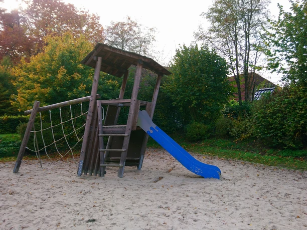 Rastede_Kinderspielplatz_Stollenkamp (2).jpg Holzspielplatz mit Sandboden, Klettergerüst, blauer Rutsche und Kletterseil umgeben von Herbstbäumen.