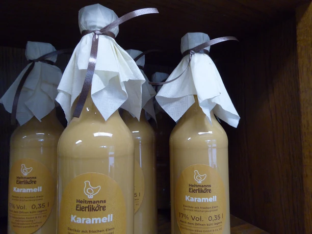 Heitmann_2.JPG Mehrere Flaschen Heitmanns Karamell-Eierlikör, dekorativ mit Schleife und Tuch auf einem Holzregal.