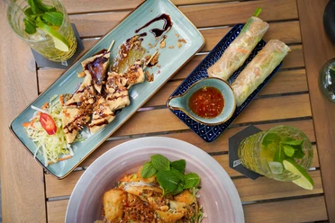 Das Bild zeigt eine Auswahl vietnamesischer Gerichte auf einem Holztisch. Links oben ist gegrilltes Fleisch mit Beilagen, rechts oben zwei Sommerrollen mit Dip; im Vordergrund eine bunte Bowl.