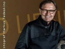 Ein Mann mittleren Alters lächelt freundlich in einem dunklen Oberteil vor einem braunen Hintergrund.