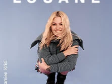 Louane in einer fröhlichen Pose vor einem blauen Hintergrund, lächelt und trägt legere Winterkleidung.