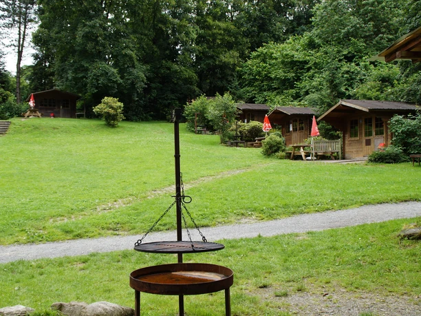 Affen- und Vogelpark Feuerstelle vor Holzhütten im Grünen, umgeben von Bäumen, mit einem geschwungenen Pfad.