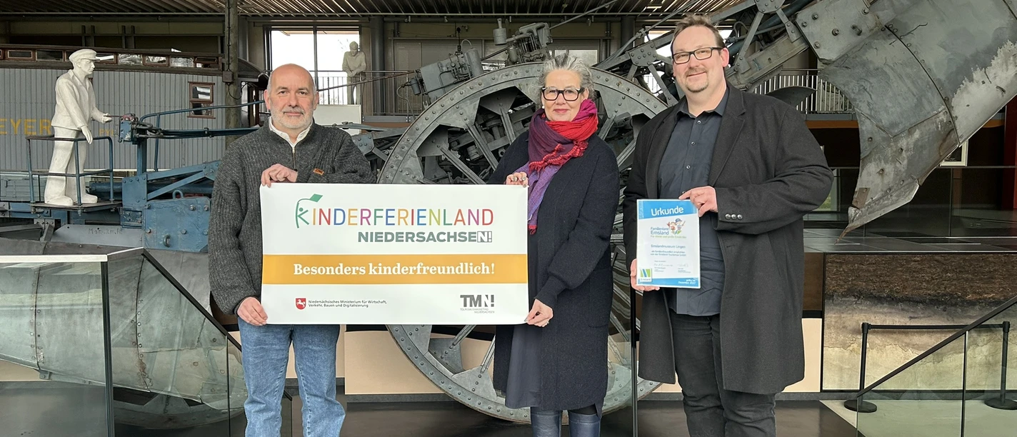 Museumsleiter mit Kinderferienland-Banner_quer ©Emsland Tourismus GmbH (1).JPEG Drei Personen stehen in einem Museum vor einer Maschine und halten ein Kinderferienland-Banner.