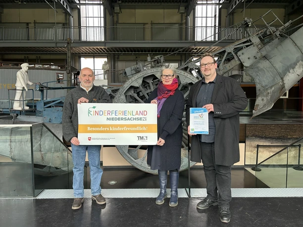 Museumsleiter mit Kinderferienland-Banner_quer ©Emsland Tourismus GmbH (1).JPEG Drei Personen stehen in einem Museum vor einer Maschine und halten ein Kinderferienland-Banner.