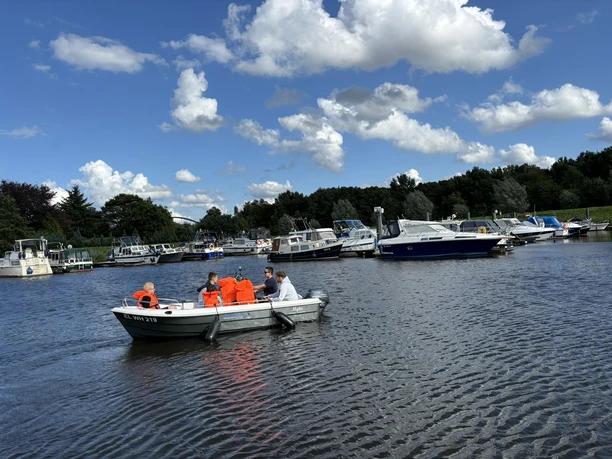 Auch Kinder lieben die Wassernähe im Emsland.JPEG Kinder mit Schwimmwesten fahren auf einem Motorboot über einen ruhigen Fluss mit anliegenden Booten.