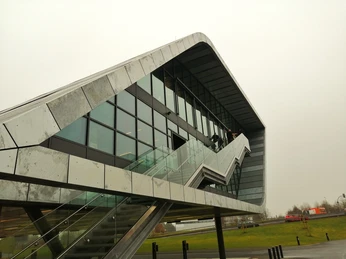 Herford-Bora Flagshipstore_Pro Herford GmbH (1).jpeg Moderne Architektur mit markantem, geschwungenem Design und großen Glasflächen bei grauem Himmel.