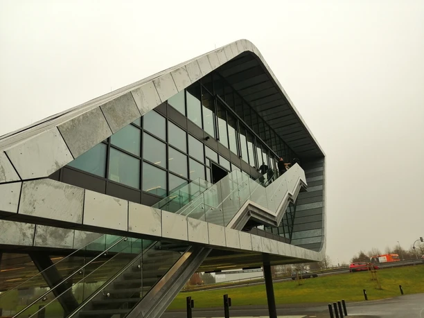 Herford-Bora Flagshipstore_Pro Herford GmbH (1).jpeg Moderne Architektur mit markantem, geschwungenem Design und großen Glasflächen bei grauem Himmel.