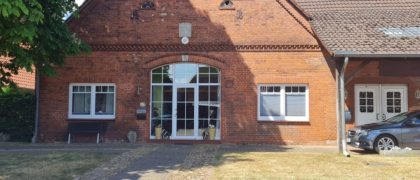 Bakstenen gebouw met grote ramen, verhard pad, een auto onder een carport aan de zijkant.