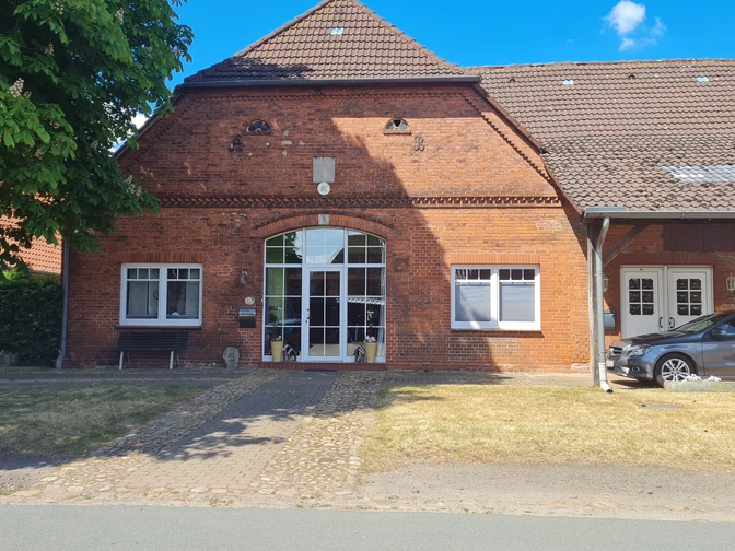 Ziegelgebäude mit großen Fenstern, gepflastertem Weg, seitlich ein Auto unter einem Carport.