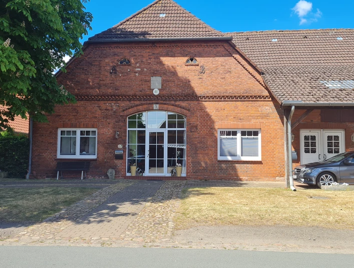 Dreppenstedt Ziegelgebäude mit großen Fenstern, gepflastertem Weg, seitlich ein Auto unter einem Carport.