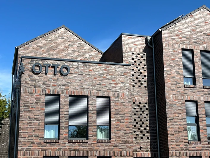 Rotes Ziegelgebäude mit modernen grauen Fensterläden und großem Schriftzug "OTTO" an der Fassade.Red brick building with modern gray shutters and large "OTTO" lettering on the façade.Rød murstensbygning med moderne grå skodder og store "OTTO"-bogstaver på facaden.Gebouw van rode baksteen met moderne grijze luiken en grote "OTTO"-letters op de gevel.