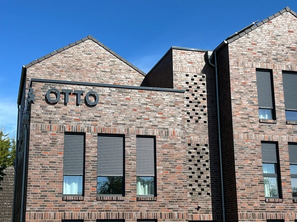 linda&otto Rotes Ziegelgebäude mit modernen grauen Fensterläden und großem Schriftzug "OTTO" an der Fassade.