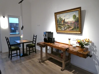 Heller Raum mit Kaffeemaschine, rustikaler Holzbank und zwei Stühlen an einem Tisch nahe einem Fenster.Bright room with coffee machine, rustic wooden bench and two chairs at a table near a window.Lyst værelse med kaffemaskine, rustik træbænk og to stole ved et bord nær et vindue.Lichte kamer met koffiezetapparaat, rustieke houten bank en twee stoelen aan een tafel bij het raam.