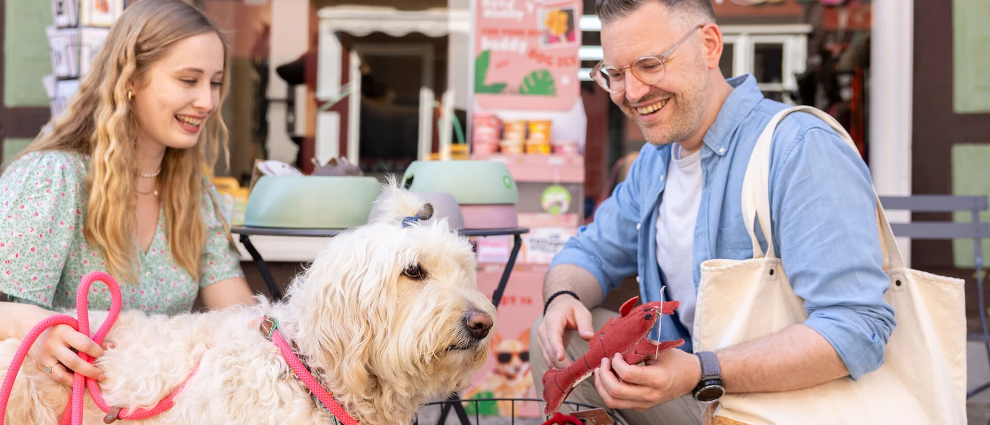 Einkaufsvergnügen in der Neuen Straße: Fein Gemacht in der Celler Altstadt Ein Paar mit einem Hund vor einem Geschäft namens "Fein Gemacht" in der Neuen Straße der Celler Altstadt. Der Mann hält ein rotes Spielzeug in Form eines Hummers, während der Hund aufmerksam zuschaut. Die Frau hält die Leine des Hundes, und beide lächeln.