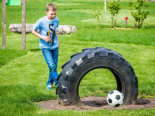 Fussballgolf Emsland - Junge kickt Ball_Hochformat ©Fussballgolf Emsland GbR.jpg Junge spielt Fußballgolf auf einer grünen Wiese und schießt den Ball durch einen großen Traktorreifen