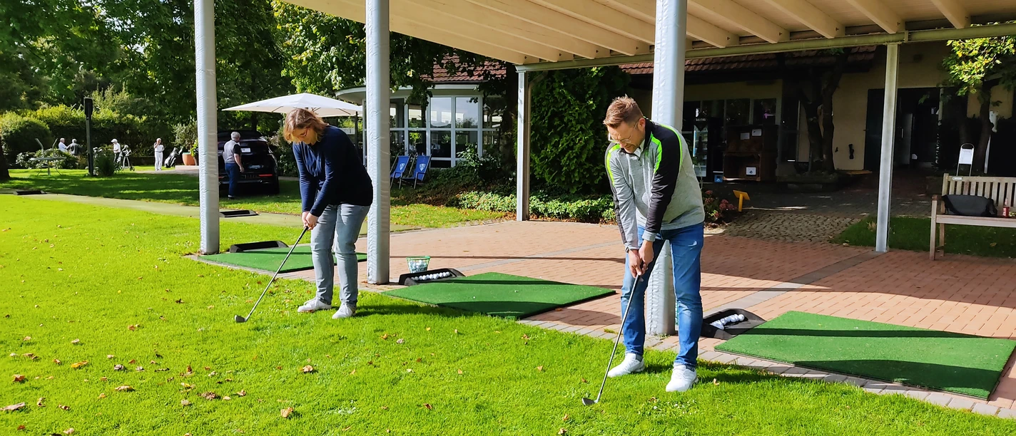 Driving Range im Golfpark Herford-IMG_20210918_111321-BloggerWG-Pro Herford GmbH.jpg Zwei Personen üben ihren Golfschwung auf der Driving Range im Golfpark Herford unter einem Vordach.