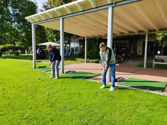 Driving Range im Golfpark Herford-IMG_20210918_111321-BloggerWG-Pro Herford GmbH.jpg Zwei Personen üben ihren Golfschwung auf der Driving Range im Golfpark Herford unter einem Vordach.