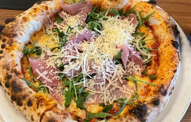 Pizza-Pro Herford GmbH.jpg Frisch gebackene Pizza mit knusprigem Rand, belegt mit Rucola, Parmaschinken und frischem Parmesan.