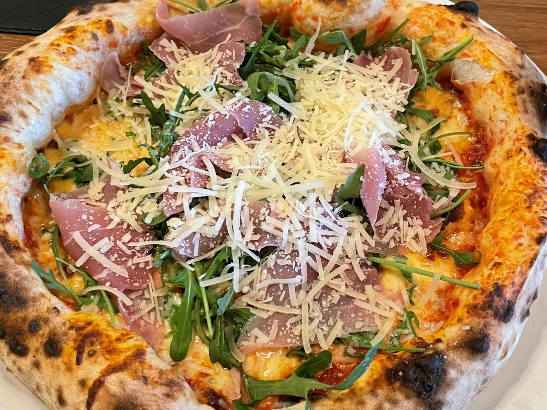 Pizza-Pro Herford GmbH.jpg Frisch gebackene Pizza mit knusprigem Rand, belegt mit Rucola, Parmaschinken und frischem Parmesan.