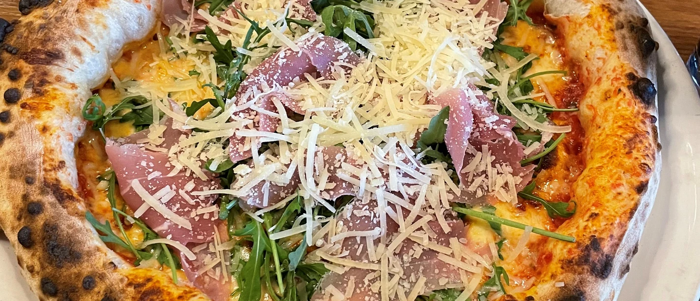 Pizza-Pro Herford GmbH.jpg Frisch gebackene Pizza mit knusprigem Rand, belegt mit Rucola, Parmaschinken und frischem Parmesan.
