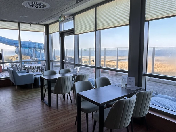 image00004.jpeg Moderner Café-Innenraum mit großen Fenstern, die einen Blick auf eine ruhige Küstenlandschaft bieten.
