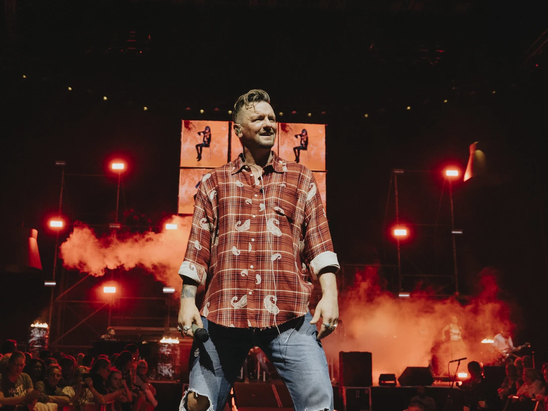 zucker_ben_chris_heidrich_.jpg Ein Sänger in kariertem Hemd performt auf einer Bühne mit roten Lichtern und rauchigem Hintergrund.