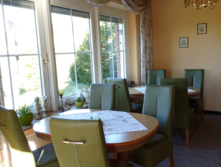 Pension Haus Marbeke Frühstücksraum.jpg Ein lichtdurchfluteter Frühstücksraum mit grünen Polsterstühlen und runden Holztischen.