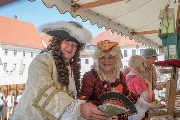 Gräfin Cosel und August der Starke dürfen in Stolpen zum Stadtfest nicht fehlen! Zwei Personen in barocken Kostümen stehen lächelnd an einem Marktstand, umgeben von historischen Gebäuden und anderen Besuchern.