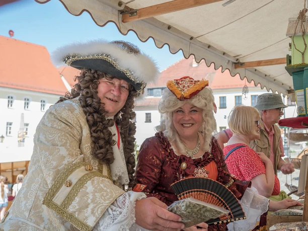 Gräfin Cosel und August der Starke dürfen in Stolpen zum Stadtfest nicht fehlen! Zwei Personen in barocken Kostümen stehen lächelnd an einem Marktstand, umgeben von historischen Gebäuden und anderen Besuchern.