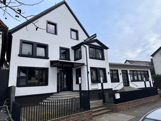 Hotel Asado- Außenansicht Hotel Asado: Weißes Gebäude mit schwarzem Dach und Fensterrahmen. Eine Treppe führt zum Hoteleingang.