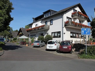 Außenansicht, weißes Gebäude mit drei Balkonen und Blumenkästen, davor befinden sich Parkplätze, links Pavillon vorm Haus mit Sitzgelegenheit