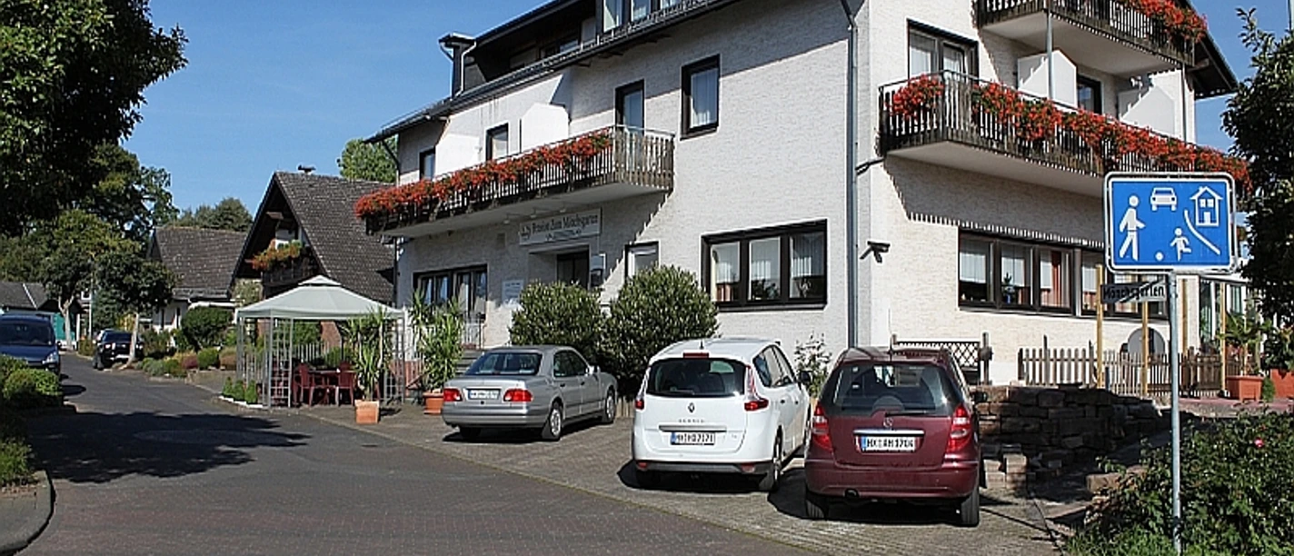 Pension Mönchsgarten Außenansicht, weißes Gebäude mit drei Balkonen und Blumenkästen, davor befinden sich Parkplätze, links Pavillon vorm Haus mit Sitzgelegenheit