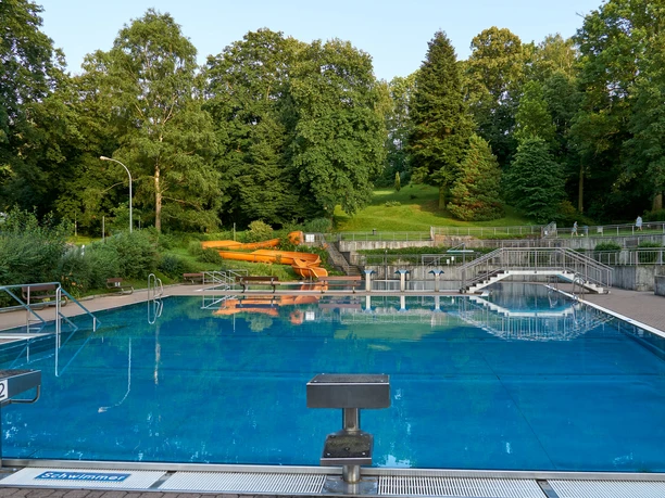 An empty swimming pool with a blue water surface, surrounded by green trees and an orange-colored slide in the background.
