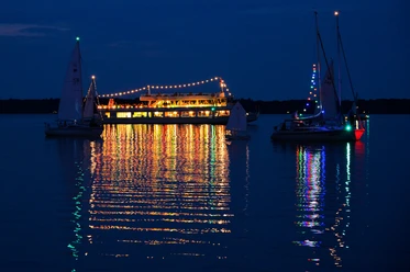 lichternacht-im-kurpark-weisse-flotte-seerundfahrt Eine festlich beleuchtete Fähre strahlt nachts auf einem ruhigen See, umgeben von reflektierenden Segelbooten.
