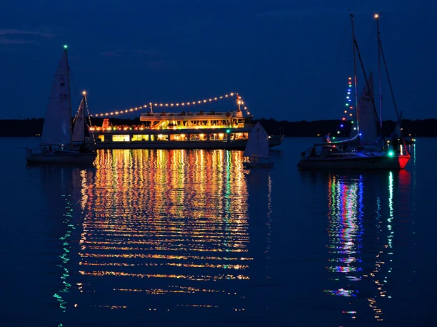 lichternacht-im-kurpark-weisse-flotte-seerundfahrt Eine festlich beleuchtete Fähre strahlt nachts auf einem ruhigen See, umgeben von reflektierenden Segelbooten.