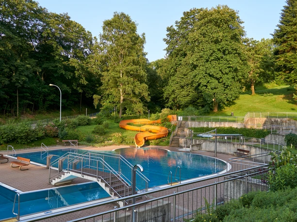 An outdoor pool with blue water, a yellow slide and surrounded by green trees and lawns in sunny weather.