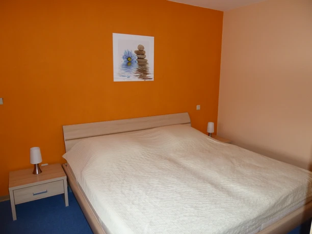 Schlafzimmer der Ferienwohnung Peter Kliesch Modernes Holz-Doppelbett, zwei Nachtschränke mit Lampen, orange Tapete im Hintergrund.