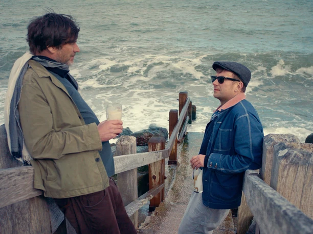 Alex James and Damon Albarn in blur To The End Ausschnitt aus dem Film "blur: To the end". Zwei Männer, jeweils mit Biergläsern in der Hand, stehen sich auf einer zum Meer führenden Treppe gegenüber.