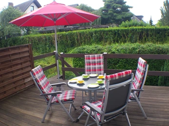 Terrasse der Ferienwohnung Udo Katzenberger Das Bild zeigt die Terrasse der Ferienwohnung Udo Katzenberger. Auf der Terrasse steht ein Tisch mit vier Stühlen, alle mit rot-weiß-karierten Polstern. Der Boden besteht aus Holz, und eine dichte grüne Hecke umgibt die Terrasse. Ein roter Sonnenschirm sorgt für Schatten und verleiht der Szenerie eine einladende Atmosphäre.
