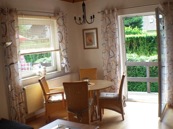 Wohnzimmer der Ferienwohnung Udo Katzenberger Ein Holz-Esstisch mit vier Korbstühlen steht vor einer geöffneten Terrassentür. Weiße Gardinen mit Muster rahmen das Fenster ein.