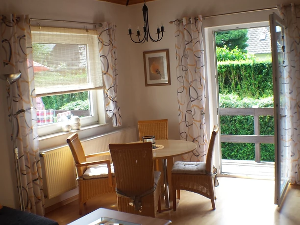 Wohnzimmer der Ferienwohnung Udo Katzenberger Ein Holz-Esstisch mit vier Korbstühlen steht vor einer geöffneten Terrassentür. Weiße Gardinen mit Muster rahmen das Fenster ein.