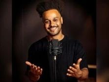 Ein lächelnder Mensch mit dunklen Locken steht vor einem Mikrofon und gestikuliert freundlich.